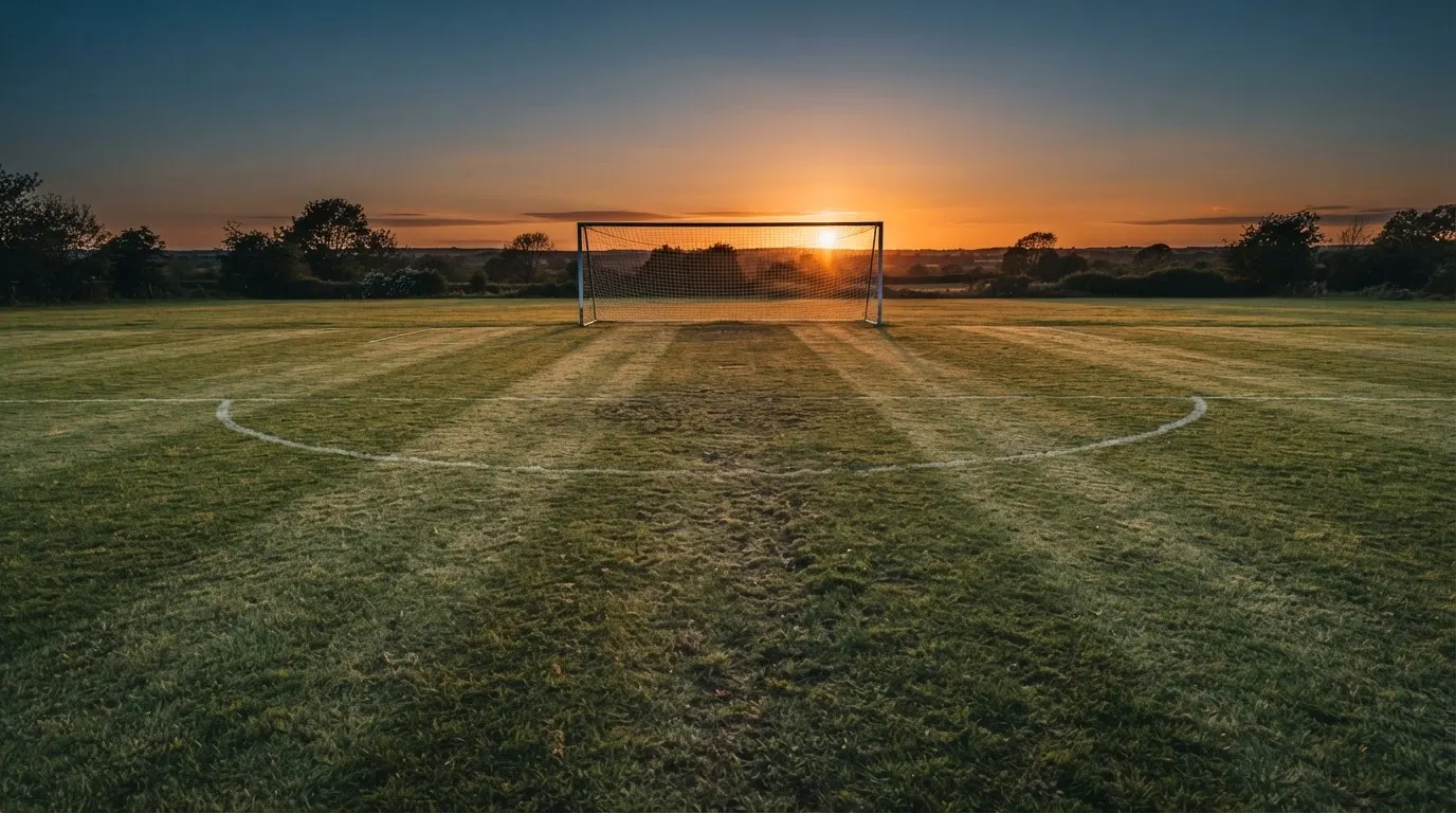 Ein Fußballtor mit Netz bei Sonnenuntergang auf einem leeren Platz, die Schatten der Torpfosten liegen lang auf dem Gras.