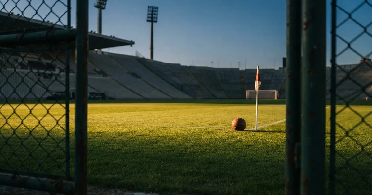 Blick durch ein Stadiontor auf einen grünen Rasen im Abendlicht, ein Ball ruht an der Eckfahne, die Tribünen sind leer.