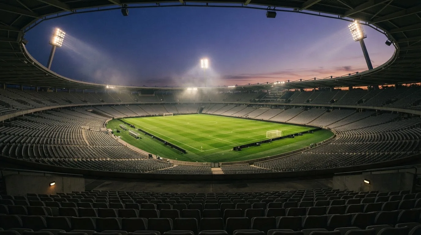Eine leere Stadiontribüne unter Flutlicht aus der Ferne betrachtet, die Rasenfläche leuchtet grün in der Dämmerung.