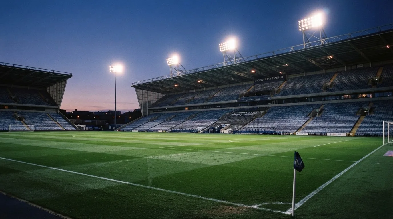 Eine leere Stadiontribüne unter Flutlicht aus der Ferne betrachtet, die Rasenfläche leuchtet grün in der Dämmerung.