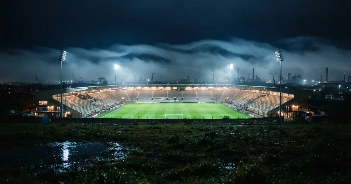 Ein Stadion unter Flutlicht, aus großer Entfernung über eine dunkle Wiese fotografiert, am Himmel zieht Nebel auf.