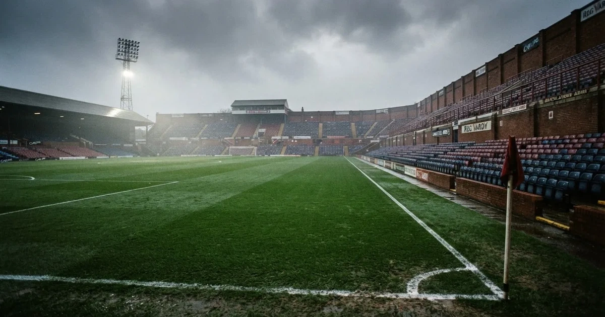 Ein englisches Fußballstadion mit rotbraunen Backsteintribünen unter bedecktem Himmel, davor ein nasser Rasen mit Spielfeldlinien.